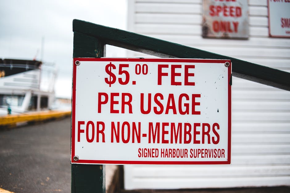 A white rectangular sign with bold red text displaying a fee of $5.00 per usage for non-members, attached to a dark green wooden frame with four metal bolts. The sign is positioned outdoors near a har